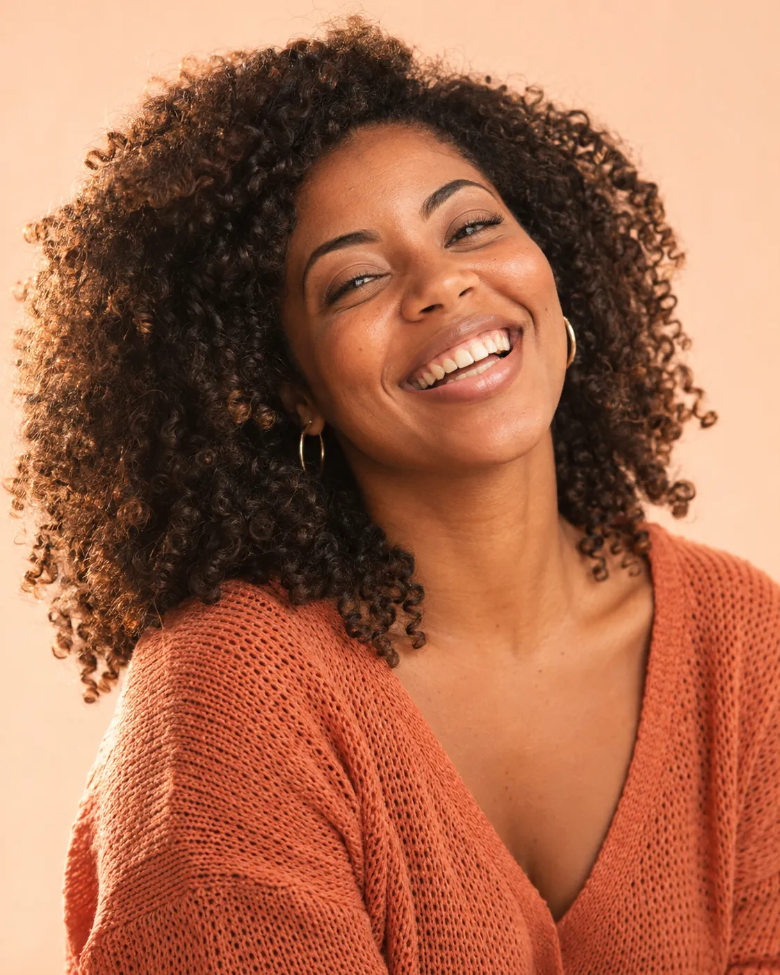 Mulher de cabelo cacheado rindo de olhos semicerrados, com a cabeça levemente inclinada, num fundo pêssego.